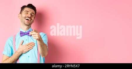 Handsome guy in bow-tie pointing finger at camera and laughing on something funny, checking out hilarious promo, standing on pin Stock Photo