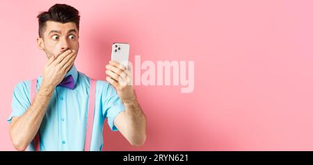 Technology concept. Silly young man looking shocked at smartphone screen while having video chat, standing on pink background an Stock Photo