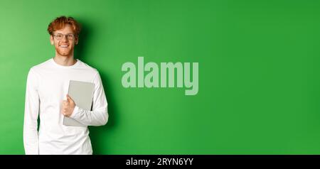 Handsome young man with red hair, wearing glasses and long-sleeve t-shirt, holding laptop on green background Stock Photo