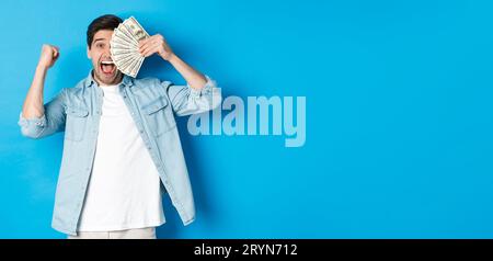 Successful young man earn money, making fist pump and showing cash, winning prize or receive credit, standing over blue backgrou Stock Photo
