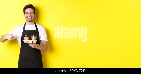 Handsome barista holding two cups of takeaway coffee, pointing finger at drinks and smiling, standing in black apron against yel Stock Photo