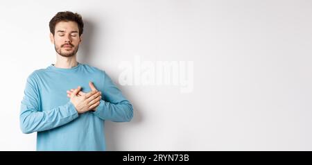 Portrait of young caucasian man with beard standing with hands on heart and closed eyes, dreaming about happy moments, standing Stock Photo