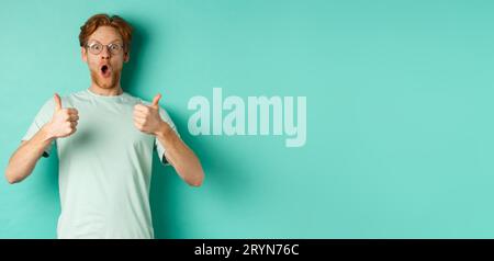 Amazed young man with red hair and beard, wearing glasses with t-shirt, showing thumbs-up and gasping in awe, checking out aweso Stock Photo