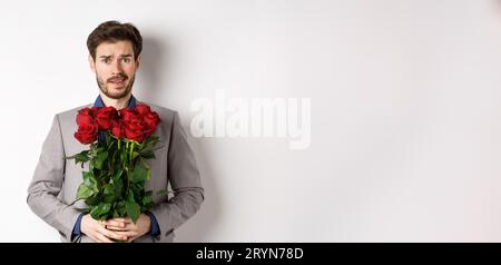 Worried boyfriend in suit, holding flowers roses and looking doubtful at camera, standing with bouquet on valentines day against Stock Photo