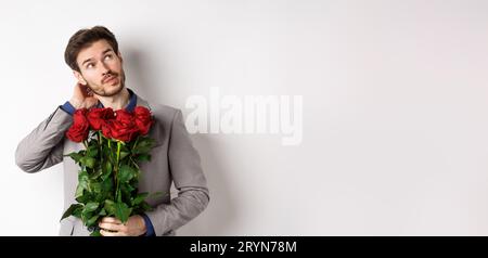 Thoughtful young man in suit going on valentines day date, holding bouquet of roses, thinking and scratching head, standing agai Stock Photo