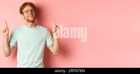 Dreamy young man with red hair and beard pointing fingers and looking cheerful up, admire something, standing over pink backgrou Stock Photo