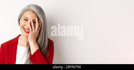 Beauty and makeup concept. Close up of beautiful asian mature woman, laughing and touching face, smiling happy, standing over wh Stock Photo