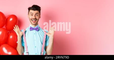 Valentines day concept. Happy young man looking surprised, raising hands up joyful, celebrating near big red hearts and pink bac Stock Photo