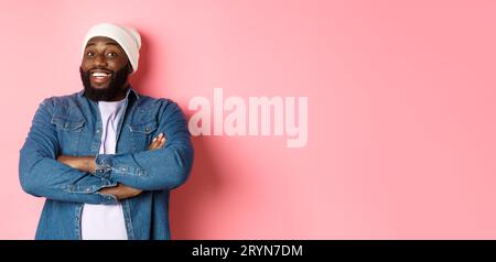 Happy Black man with beard, wearing beanie and denim shirt, looking intrigued and amused at camera, smiling with arms crossed on Stock Photo