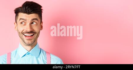 Close-up of cheerful smiling man, looking aside at logo, stare at promo deal with excited face, standing on pink background Stock Photo