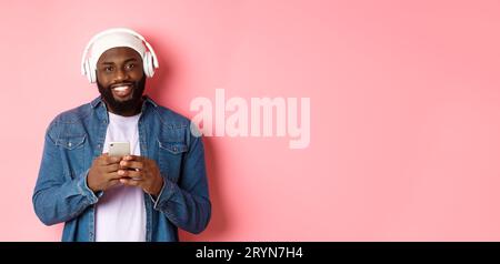 Image of handsome Black guy in headphones, listening music and using mobile phone, smiling at camera, pink background Stock Photo