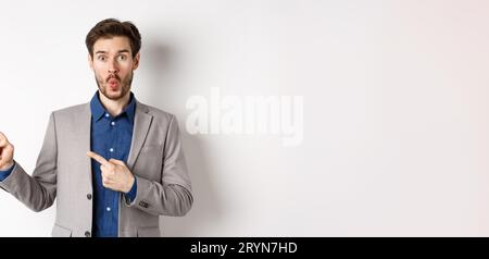 Excited handsome man in suit pointing fingers left at banner and saying wow, look here gesture, showing advertisement, standing Stock Photo