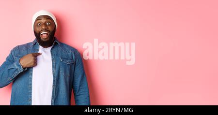 Surprised Black man in hipster beanie pointing at himself, staring at camera amazed, standing over pink background Stock Photo