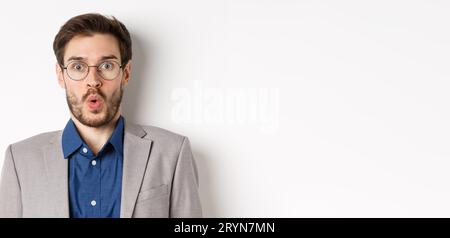 Close up portrait of excited office worker in glasses and suit saying wow, staring amazed at camera, standing against white back Stock Photo