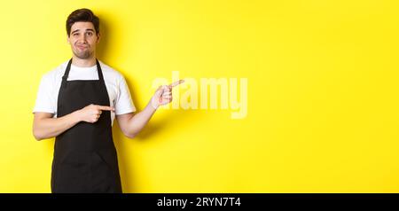 Doubtful barista in black apron pointing fingers right, looking skeptical and unamused, standing over yellow background Stock Photo