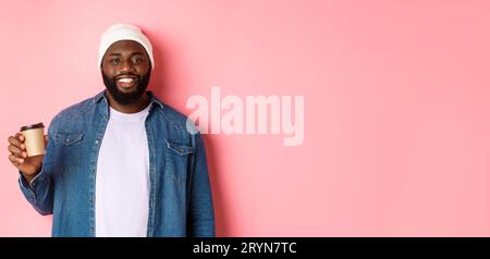 Handsome modern Black man drinking takeaway coffee, smiling and looking satisfied at camera, standing over pink background Stock Photo