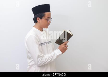 Portrait of young Asian muslim man reading and reciting Holy book of Quran seriously. Isolated image on white background Stock Photo