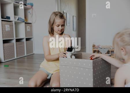 Children cleaning toys from floor after playing, collect them to textile box. Kids sitting on carpet at white home bedroom, kindergarten playroom or p Stock Photo