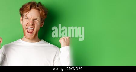 Close-up of lucky young man winner, celebrating victory, shouting yes with joy and making fist pumps, triumphing on green backgr Stock Photo