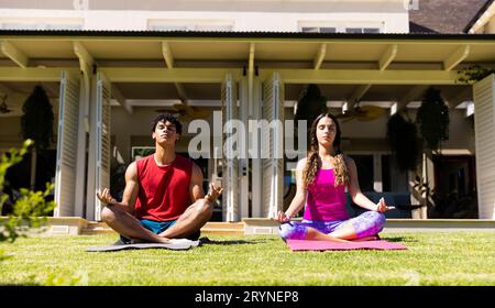 Biracial young couple practicing meditation while sitting on mats in yard against house Stock Photo