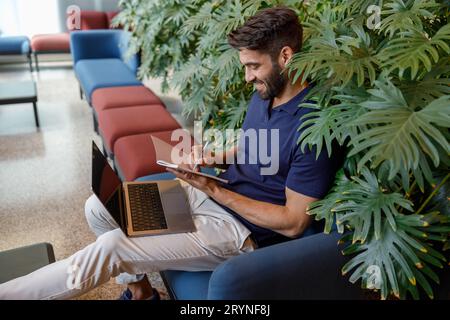 Handsome smiling freelancer is making notes during work on laptop in office hall Stock Photo