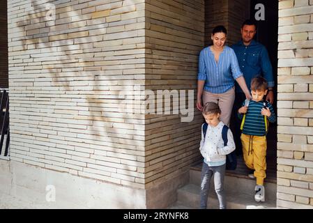 Young parents seeing their cute kids off to school. Smiling mom, dad, daughter and son go down the steps of the terrace of their Stock Photo