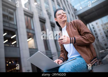 Smart attitude. Positive handsome man using a laptop and sitting in the street while surfing the internet have a web conference. Stock Photo