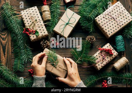 Top view of a wooden surface strewn with spruce branches and gifts. Female hands are holding one gift box. Stock Photo
