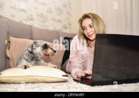 Woman is lying on the carpet at home with a laptop and looking at her cute gray dog who is asking for attention Stock Photo