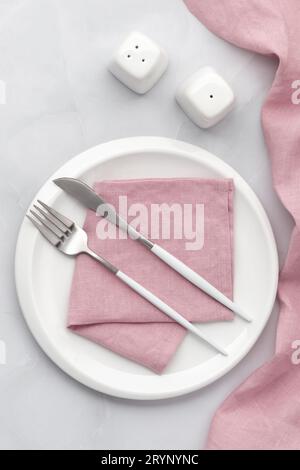 White plate with silver cutlery. Knife, fork and pink napkin on grey background. Table setting, flat lay, top view. Serving space, clean ceramic dish. Stock Photo
