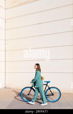 Young empowered ginger-haired woman in green suit walking with bicycle at the financial district with modern building on the background Stock Photo