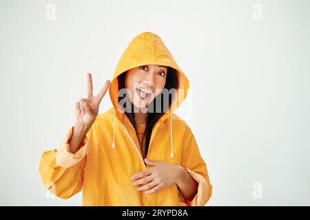 Joyful asian girl with braces dressed in bright yellow raincoat showing sign of victory and peace on white studio background Stock Photo