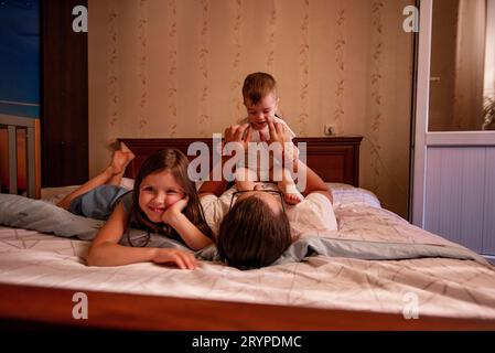 Diversified father in glasses throws baby son on the bed, eldest daughter is having fun nearby. Family fools around and plays on weekends. Father's da Stock Photo