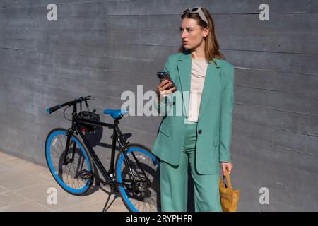 Young empowered ginger-haired woman in green suit using smartphone standing with her bike on black background Stock Photo