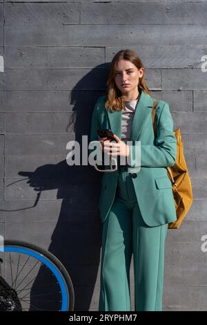 Stylish business ginger-haired woman in green suit and yellow backpack standing with bicycle at black background Stock Photo