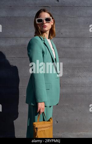 Stylish business ginger-haired woman in green suit, white sunglasses and yellow backpack standing on black background Stock Photo