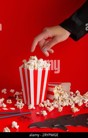 Womans hand taking popcorn from striped box, bat napkin and straws on a red background. Stock Photo