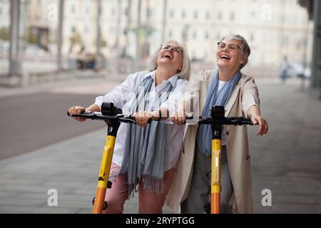 Portrait happy laughing Senior women with gray hair enjoying ride together on electric scooters outdoors. Two retired female friends at city street. Stock Photo