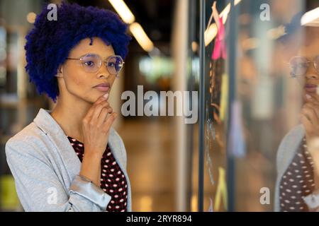 Thoughtful biracial casual businesswoman with blue afro brainstorming at glass wall in office Stock Photo