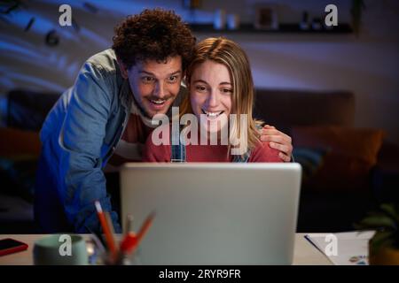 Surprised Caucasian couple looking happy at computer sitting at desk in dark living room.  Stock Photo