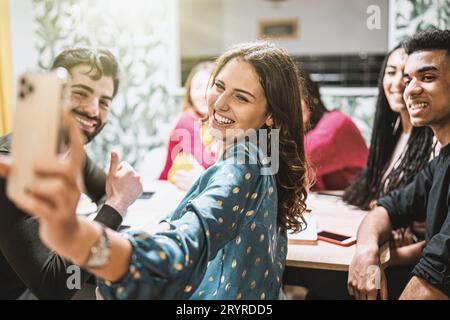 A diverse group of friends gathers around the kitchen table, smiling as a young woman takes a group selfie with her phone. Stock Photo