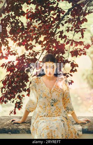 A young woman sitting alone in a park under a tree a relaxed pose, wearing a stylish dress Stock Photo