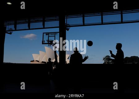 Two men in silhouette tossing a round ball with the Sydney Opera House framed by the silhouette of the OSPT cargo hall & an American Basketball hoop Stock Photo