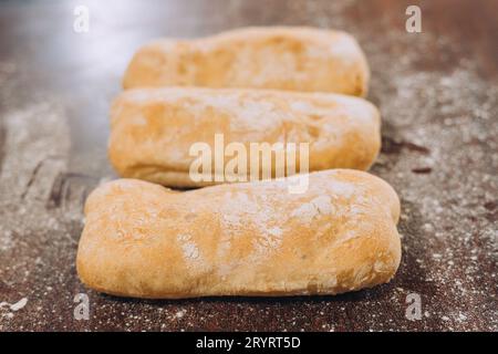 Three golden-brown loaves of freshly-baked bread sitting on a wooden table in a rustic kitchen setting Stock Photo