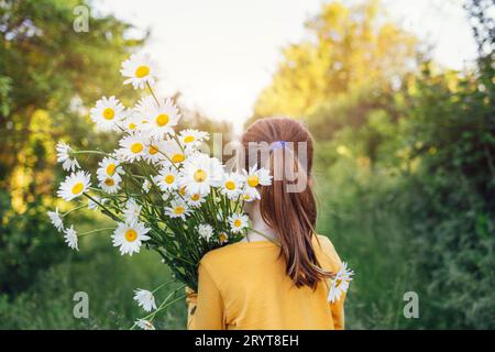 Little girl in yellow t-shirt carries huge bouquet of white flowers on her shoulder outdoors on meadow. Stock Photo