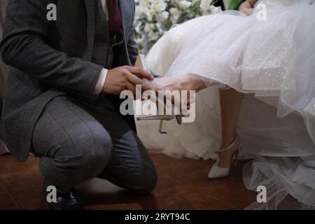 A groom in formal attire kneels while cutting a three-tiered white wedding cake, ready to share the first slice with his bride Stock Photo