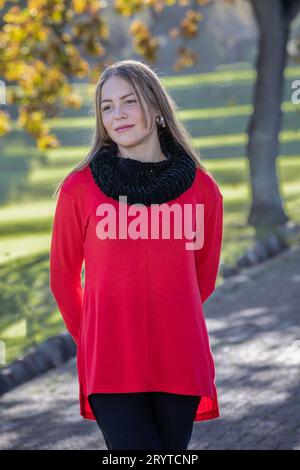 In a head and shoulders shot, a stunning young blonde woman, wearing a black woolen hat and a bright red sweater, radiates under the autumn trees Stock Photo