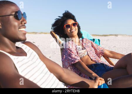 Happy african american couple sitting in deckchairs holding hands on sunny beach Stock Photo