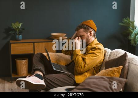 Sad and tired man is depicted sitting on a sofa at home. Overwhelmed by bad news or challenges, he holds his head with closed ey Stock Photo