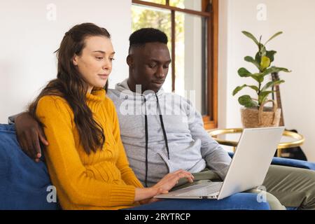 Diverse couple sitting on sofa using laptop at home Stock Photo
